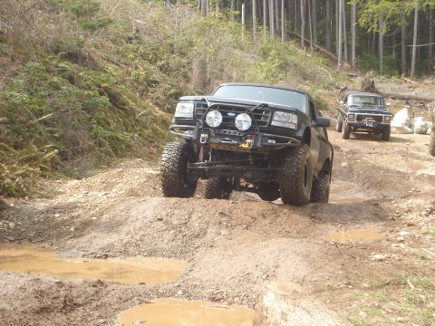 1989 Ford Bronco - Modified 1989 Ford Bronco tackles a challenging dirt path in a wooded area, with a second vehicle in tow.