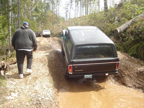 1989 Ford Bronco - A black Ford Bronco navigates a muddy path on a forest trail, with other vehicles visible ahead.