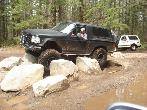 1989 Ford Bronco - A black Ford Bronco is shown off-roading over large rocks, with another white Bronco visible in the background.