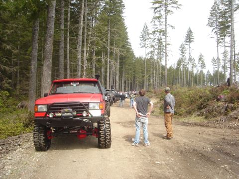 1989 Ford Bronco - Several off-road vehicles line a dirt path through a forest with people standing around.