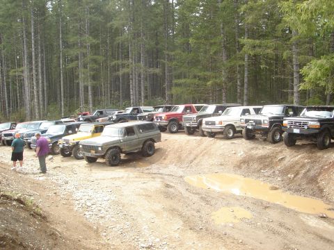 1989 Ford Bronco - Multiple SUVs and trucks are parked on a dirt track in a wooded area, with a muddy puddle in the foreground.