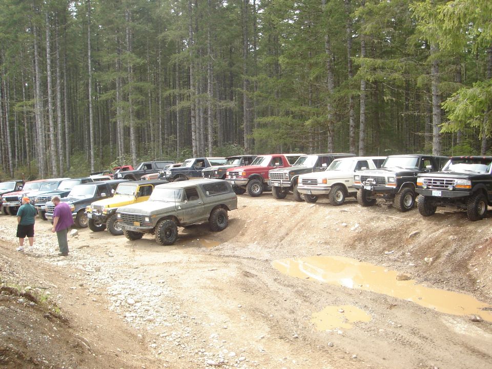 Line of off-road vehicles parked on a dirt track with trees in the background. The lead vehicle is a dark green SUV