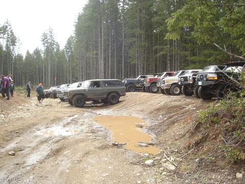 1989 Ford Bronco - Several off-road vehicles are lined up on a dirt track next to a dense forest, with a muddy puddle in the foreground.