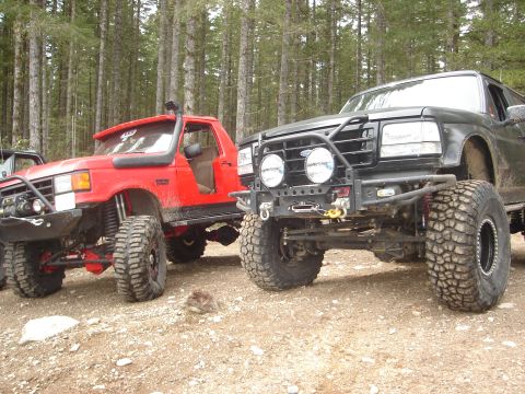 1989 Ford Bronco - Two off-road trucks, a red one and a black one, are parked side-by-side on a gravelly path surrounded by trees.