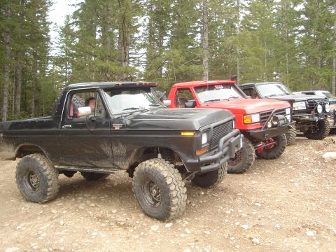 1989 Ford Bronco - A line of off-road vehicles, including a black SUV with large tires, parked on a dirt road with trees in the background.