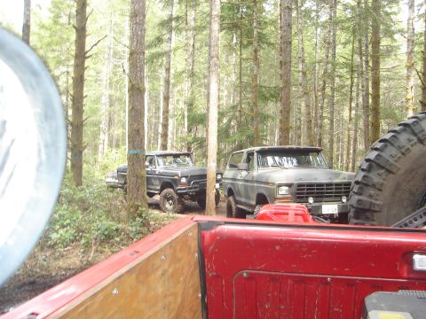 1989 Ford Bronco - Two off-road vehicles are parked on a trail surrounded by tall trees in a wooded area.