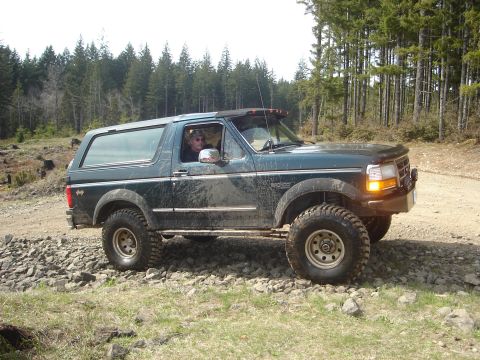1989 Ford Bronco - This dark green Ford Bronco is shown off-road on a rocky path, with trees in the background.