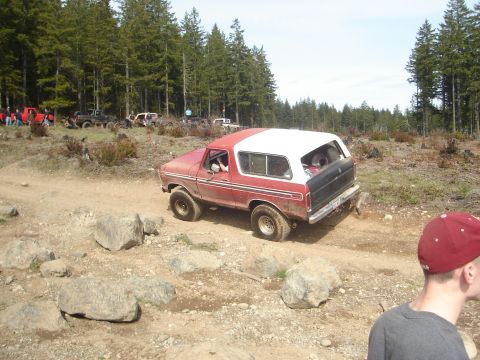 1989 Ford Bronco - A red and white Ford Bronco navigates a rough, wooded trail, with other vehicles visible in the distance.