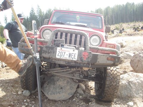 1989 Ford Bronco - A red Jeep Wrangler is stuck on a large rock while off-roading, with a hi-lift jack being operated by a person.