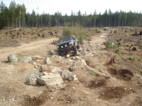 1989 Ford Bronco - A black 1989 Ford Bronco tackles a challenging rocky terrain on an off-road trail, with another vehicle visible in the distance.