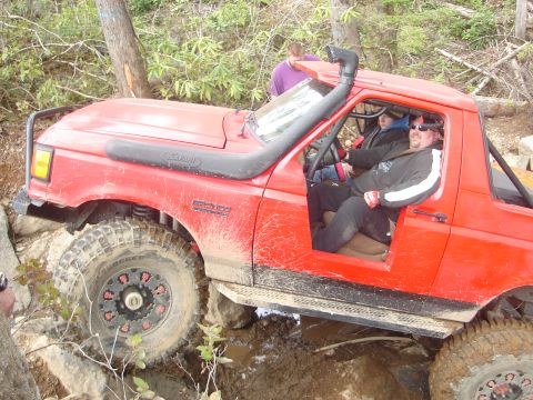1989 Ford Bronco - Red 1989 Ford Bronco off-roading through mud and rocks with occupants inside.