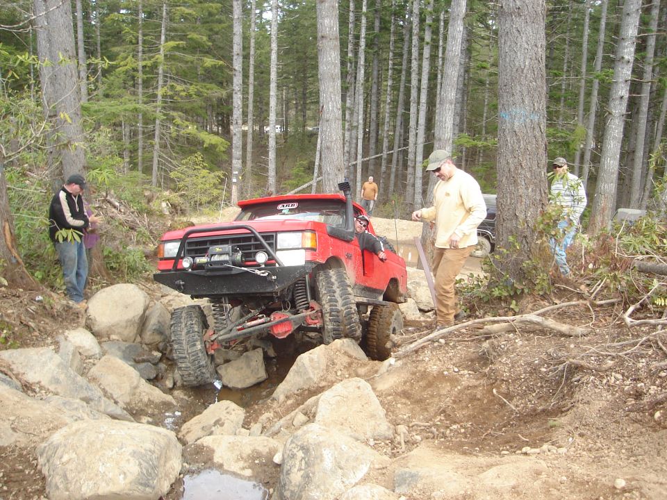 red off-road truck going over rocks, guy in tan shirt watching