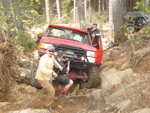 1989 Ford Bronco - A red 1989 Ford Bronco is being winched through a challenging rocky trail with assistance from people and another vehicle.