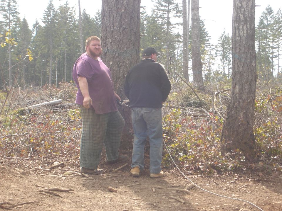 Two men stand in a wooded area with trees and brush