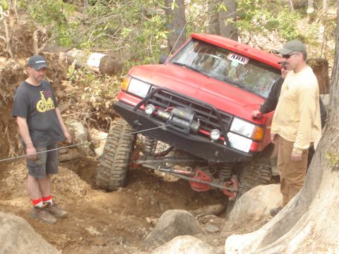 1989 Ford Bronco - A red off-road vehicle is being winched through a challenging rocky obstacle course with onlookers present.