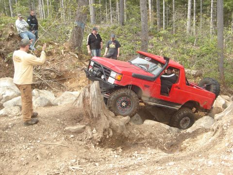 1989 Ford Bronco - A red off-road vehicle navigates a challenging obstacle on a wooded trail with spectators observing.