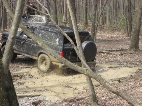 1991 Ford Bronco - Two vehicles navigate a muddy trail through a forest, with one appearing to be stuck.