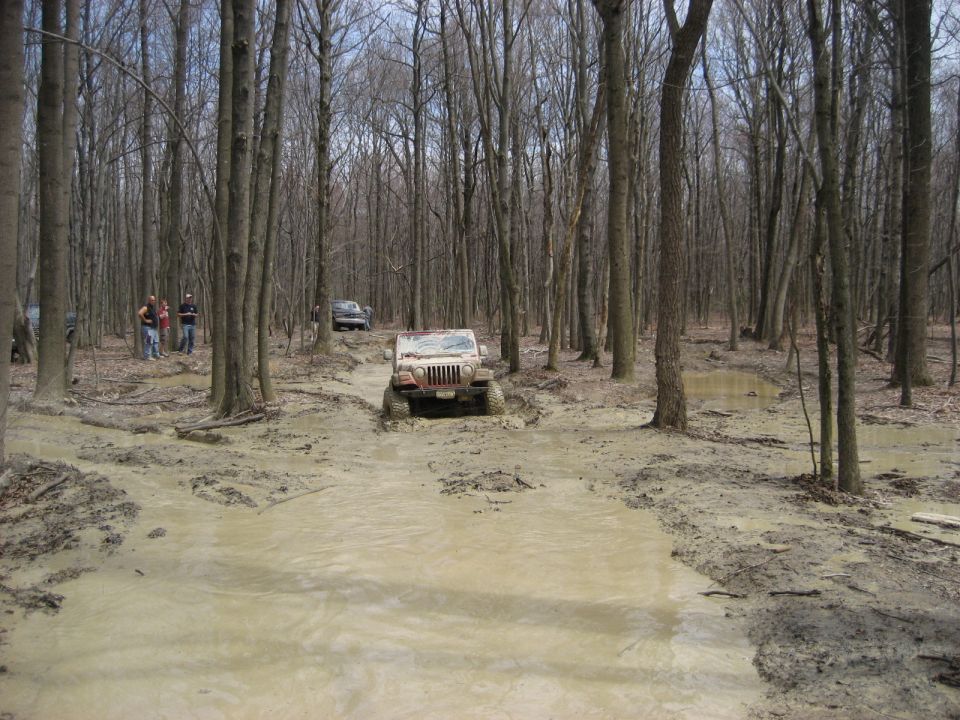 Muddy red Jeep Wrangler crossing a flooded rutted trail in bare woods with onlookers