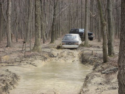 1991 Ford Bronco - Two trucks are shown driving through a muddy path in a forest during the day.