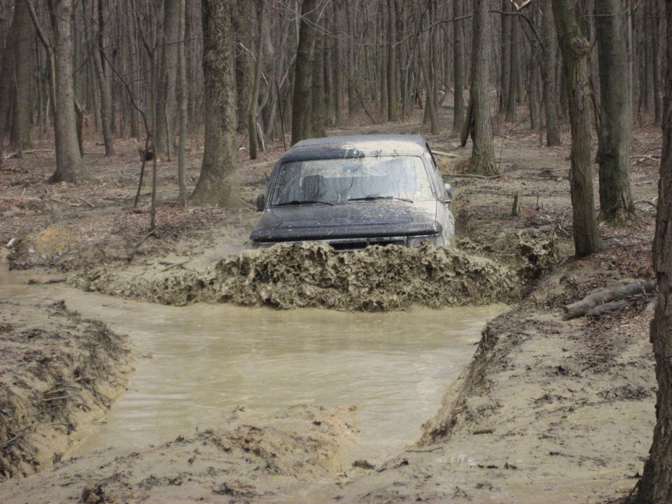 Dark truck buried to the hood pushing a wall of mud through a deep water hole in the woods