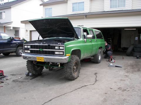 1990 Chevrolet Suburban - A green 1990 Chevrolet Suburban is parked outside a garage with its hood up, undergoing maintenance.