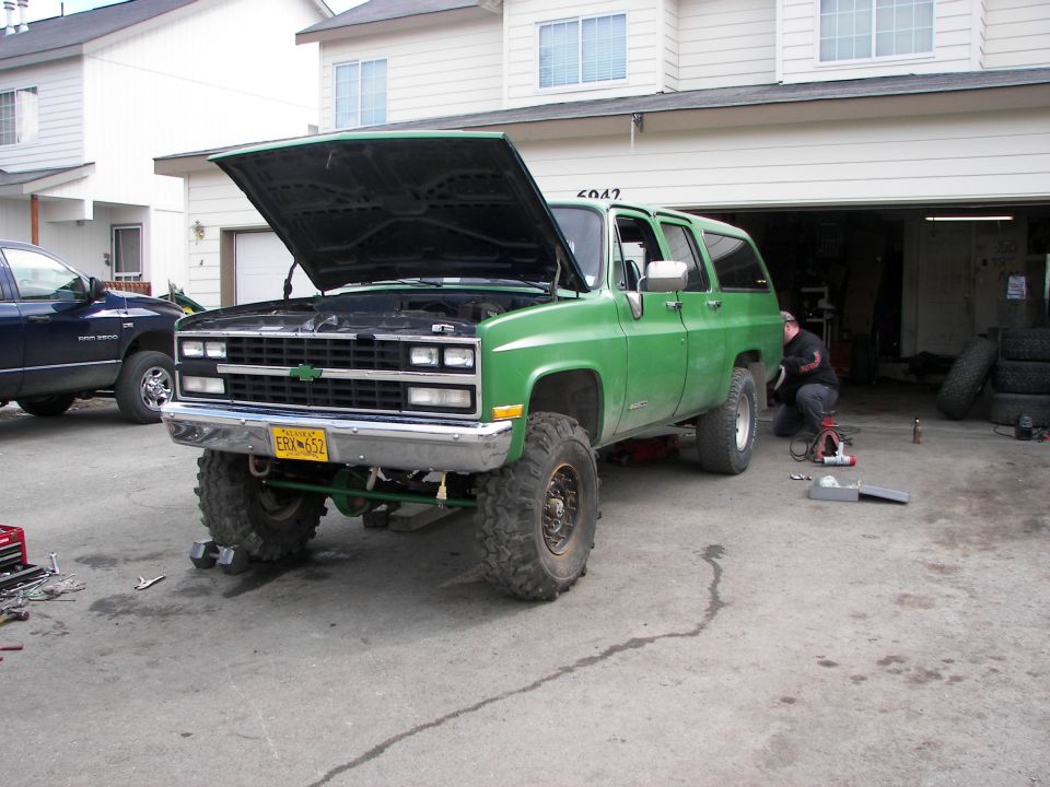 Green 1990 Chevrolet Suburban with hood open, lifted on large tires, parked in driveway