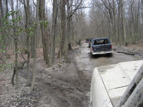 1991 Ford Bronco - A convoy of Ford Broncos navigates a challenging muddy path through a dense forest.