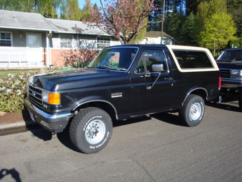 1991 Ford Bronco - A black 1991 Ford Bronco with a tan removable top is parked on a street next to another vehicle.