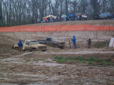 1979 Ford F-250 - Two pickup trucks are stuck in a mud pit during an event, with spectators watching from a distance.