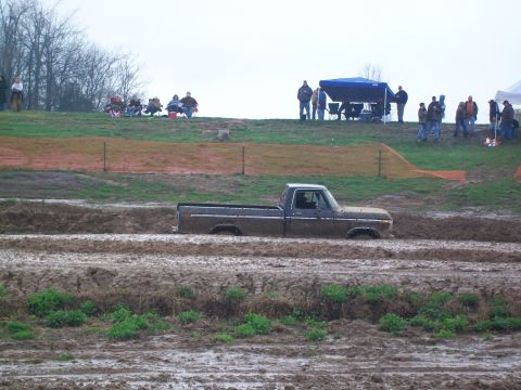 1979 Ford F-250 - A dark-colored pickup truck is partially submerged in a mud track, with onlookers gathered on a grassy hill in the background.