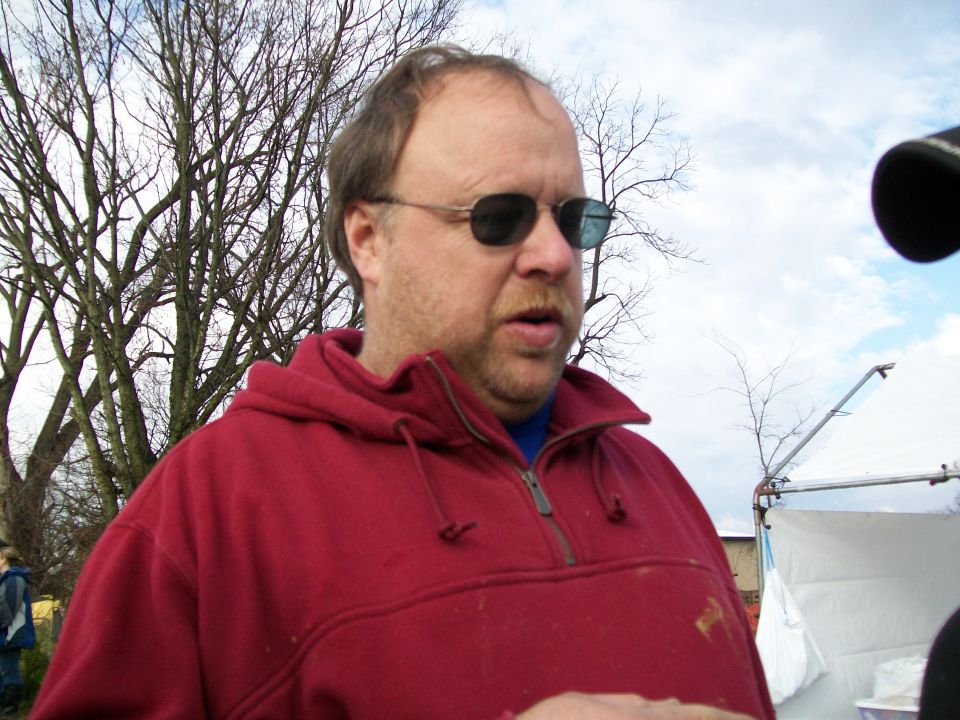 Man wearing sunglasses and a red hoodie, talking outdoors with trees and a tent in the background