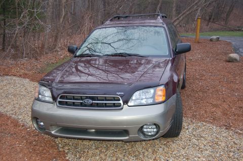 2001 Ford Crown Victoria - A maroon Subaru Outback with tan lower body panels is parked on a gravel surface near trees.