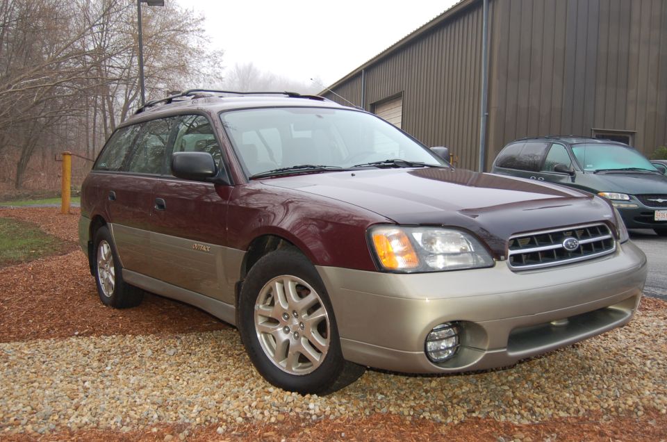 Maroon and tan Subaru Outback Wagon parked on gravel. Another dark SUV is visible in the background