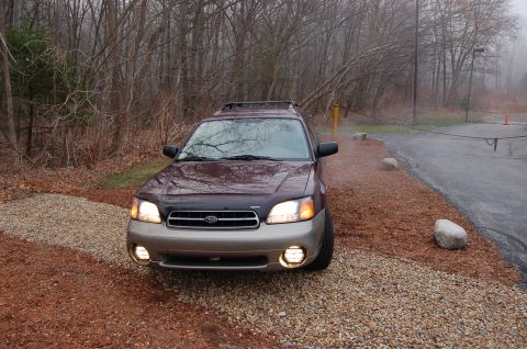 2001 Ford Crown Victoria - A maroon and tan Subaru Outback wagon is parked on a gravel area next to a paved road.
