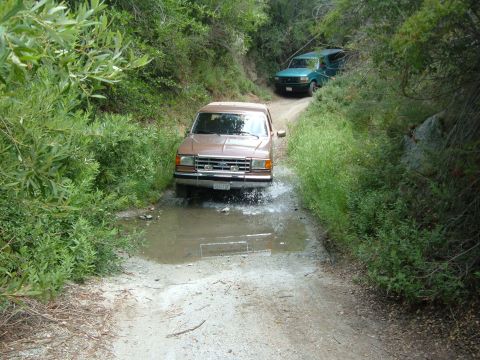 1988 Ford Bronco - A copper/tan Ford Bronco splashes through a shallow water crossing on a narrow gravel trail hemmed in by green brush, front end low with sp…