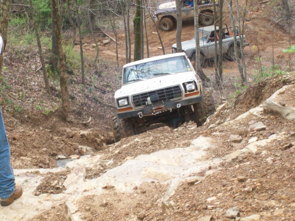White lifted Ford 4x4 with winch bumper climbing a rocky off-road ledge in the woods