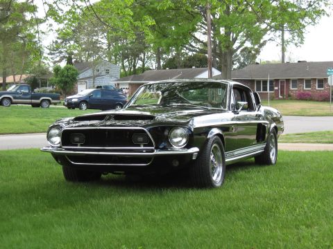 1968 Shelby GT500 - A black 1968 Shelby GT500 with racing stripes is parked on a grassy lawn in front of a suburban house.