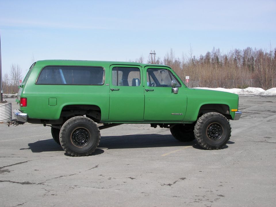 Green lifted Chevrolet Suburban parked on asphalt with trees and snow in background
