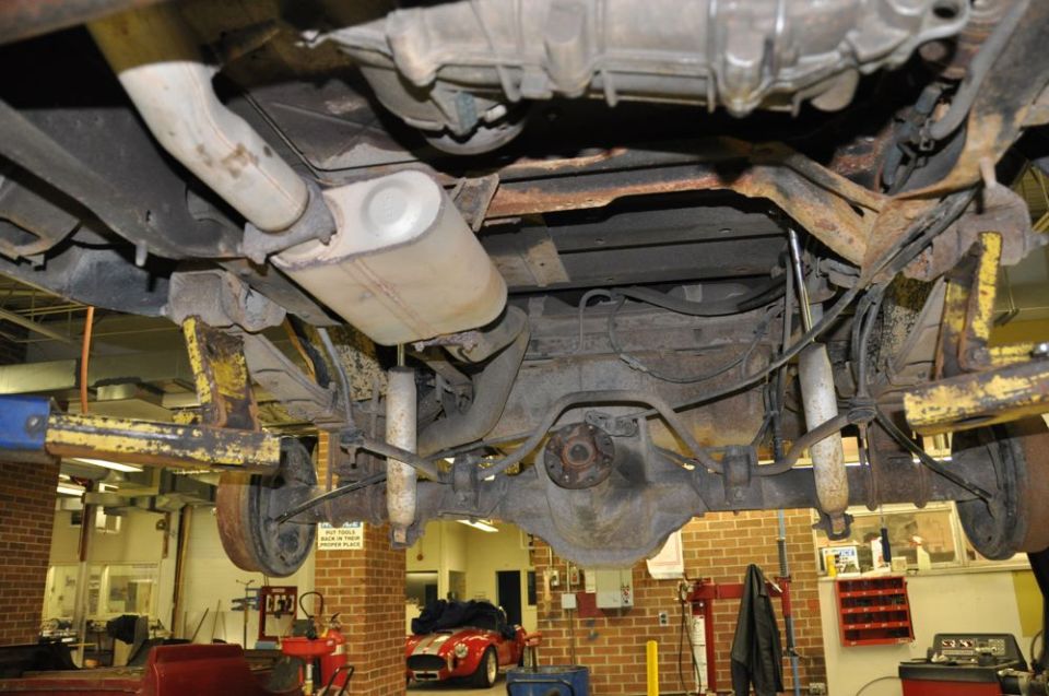Undercarriage view of a vehicle on a lift, showing suspension and exhaust components. A red sports car is visible in the background