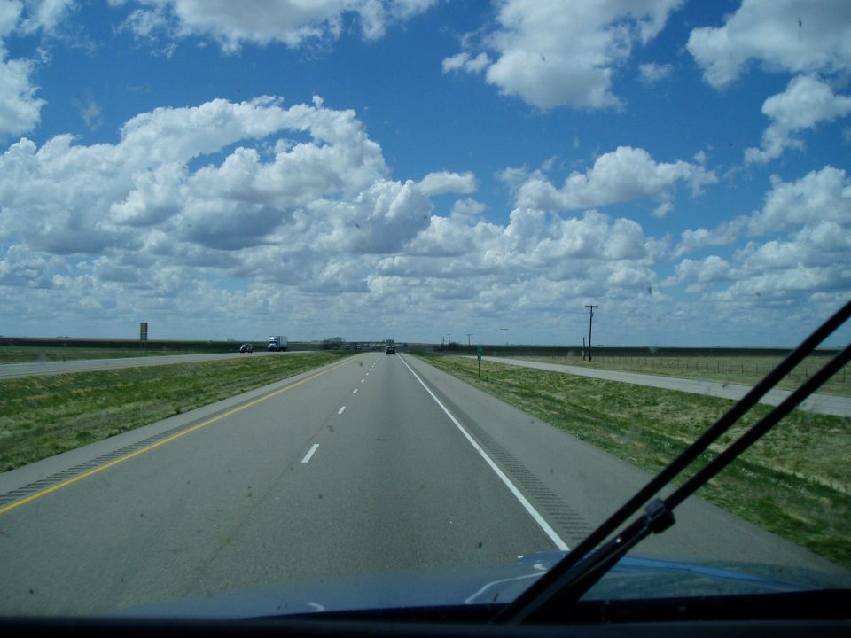 Windshield view of a divided highway through flat grassland under blue cloudy sky.