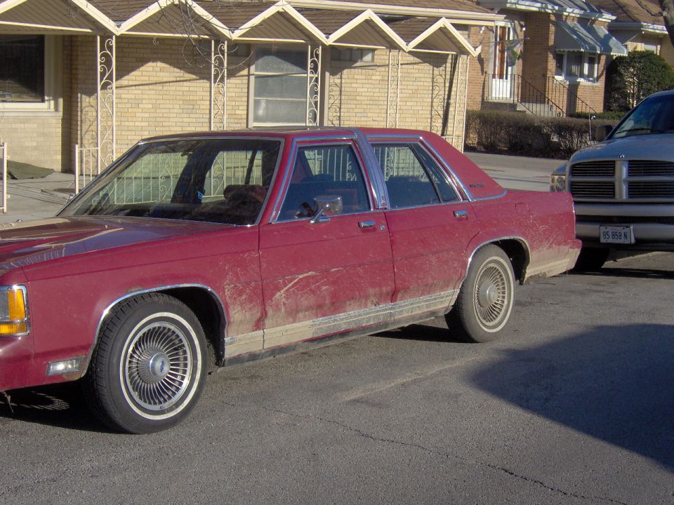 Dirty dark red Ford LTD Crown Victoria sedan, driver side view, parked on street with a Dodge pickup behind it