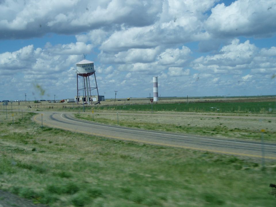 Leaning 'Britten USA' water tower beside a rural highway under cloudy sky