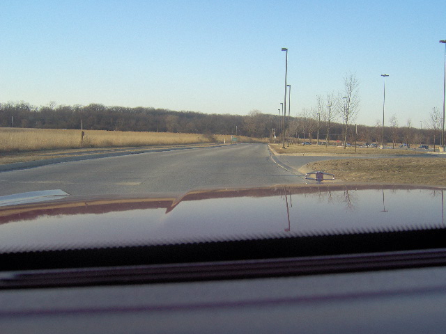 View from inside a car looking out at a road, fields, and trees under a clear sky