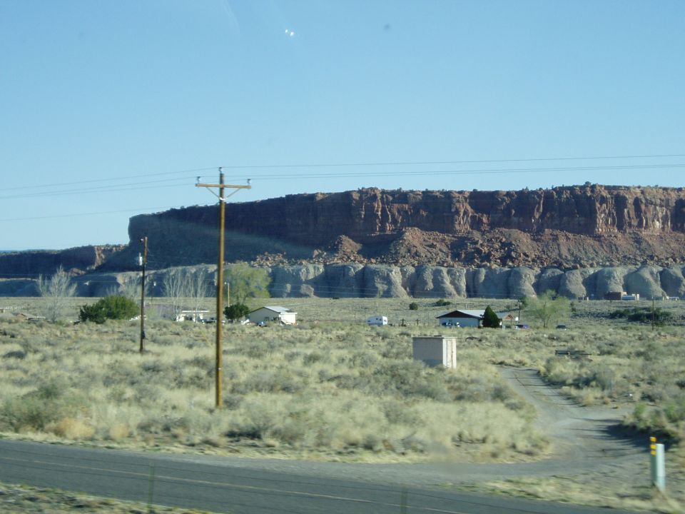 Desert landscape with mesas, houses, and telephone poles under a clear blue sky. A dirt road leads to a house