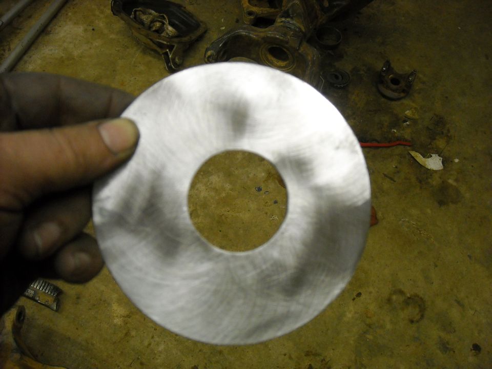 Hand holding a circular metal part with a hole in the center. Background shows a concrete floor with some mechanical parts