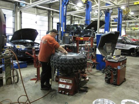 1992 Ford Bronco - A mechanic works on a large tire at a tire changing machine in a garage, with other vehicles visible in the background.