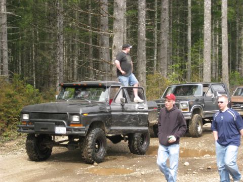 1979 Ford Bronco - Several off-road vehicles are parked on a dirt path surrounded by tall trees.