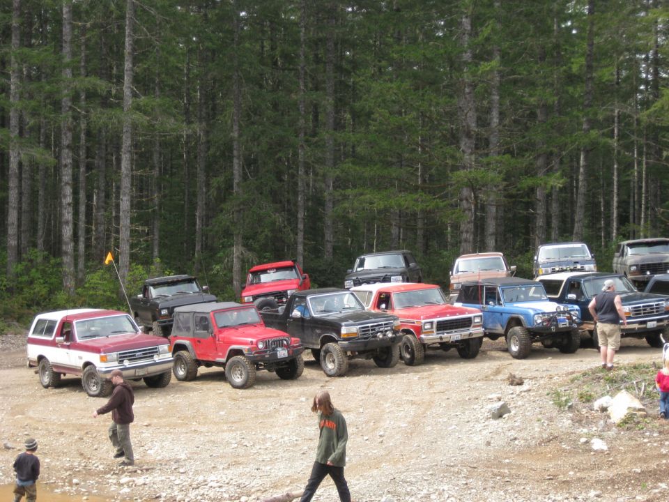 Several off-road trucks and SUVs parked in a dirt lot with a forest background. Some people are standing near the vehicles