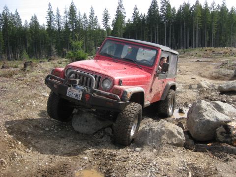 1979 Ford Bronco - A red Jeep Wrangler tackles a challenging off-road course, with its tires positioned on large rocks.