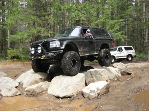 1979 Ford Bronco - Modified black Ford Bronco driving over rocks on a trail, with another Bronco behind it.